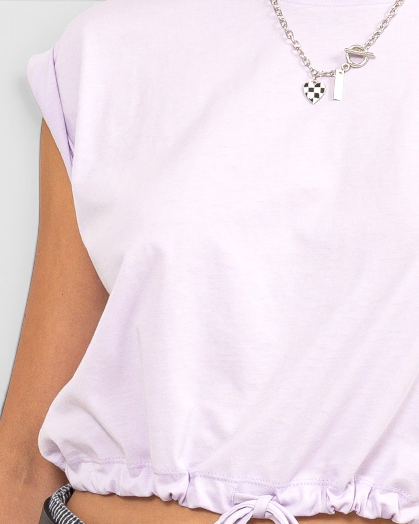 A close-up of a person wearing a light lavender sleeveless top with a drawstring hem, paired with a silver necklace featuring a checkered heart and the number 1.