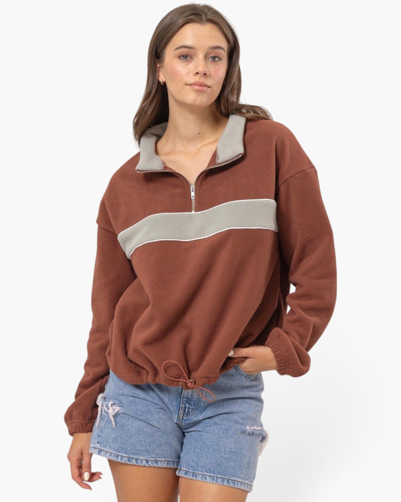 A woman modeling a rust-brown half-zip sweatshirt with a contrasting light gray collar and horizontal chest stripe, paired with light-wash denim shorts against a plain white background.