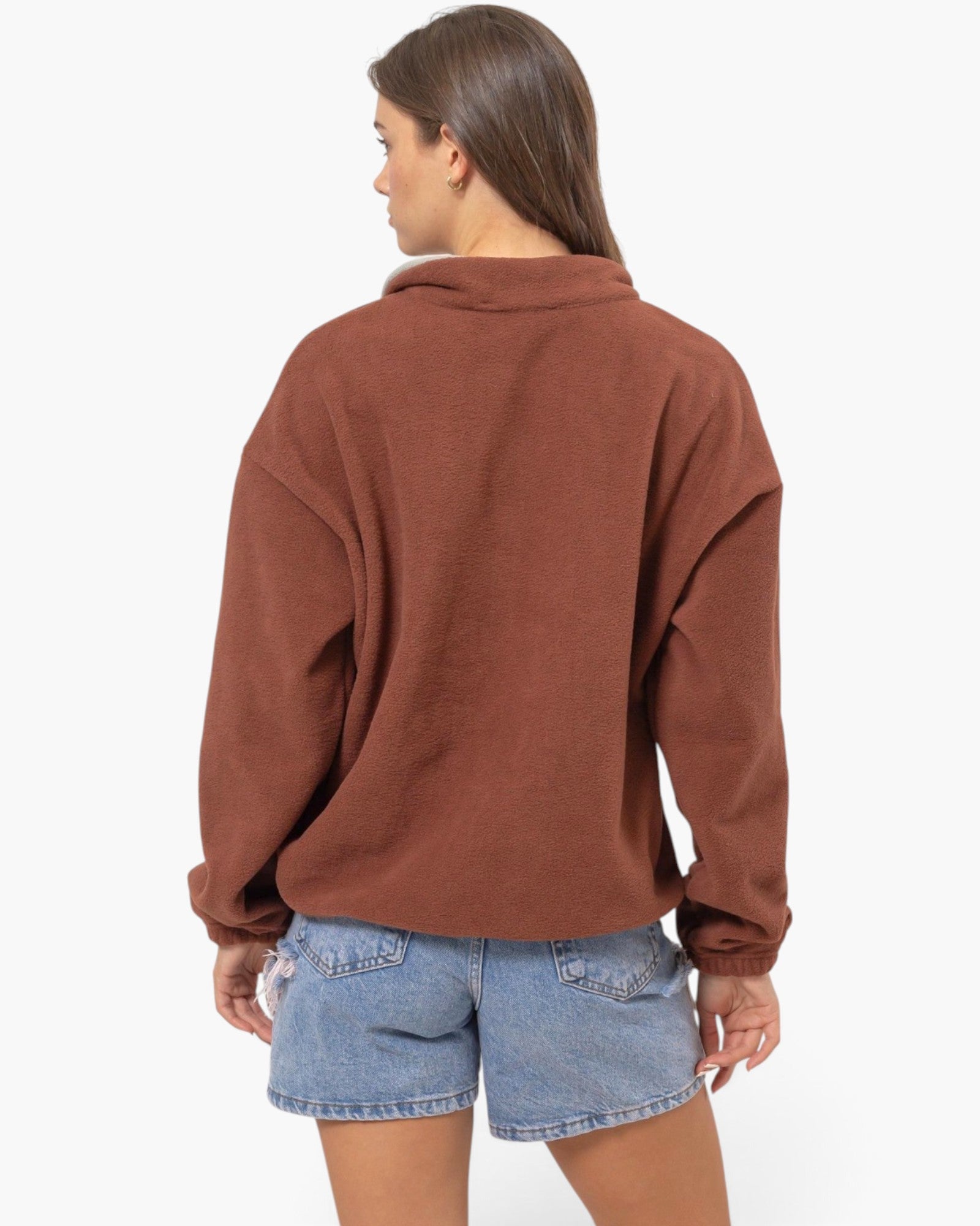A woman shown from the back wearing a rust-brown oversized pullover sweatshirt and light-wash denim shorts against a plain white studio background.