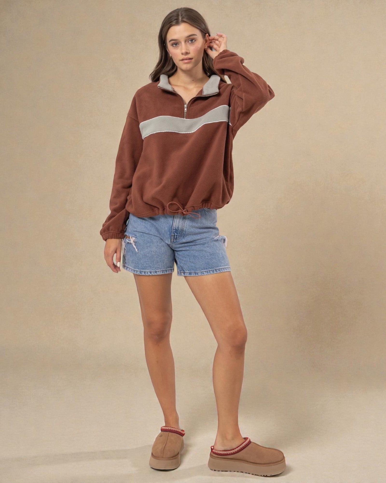 Woman wearing a brown sweatshirt with a white stripe, blue shorts, and brown shoes on a beige background
