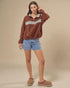 Woman wearing a brown sweatshirt with a white stripe, blue shorts, and brown shoes on a beige background