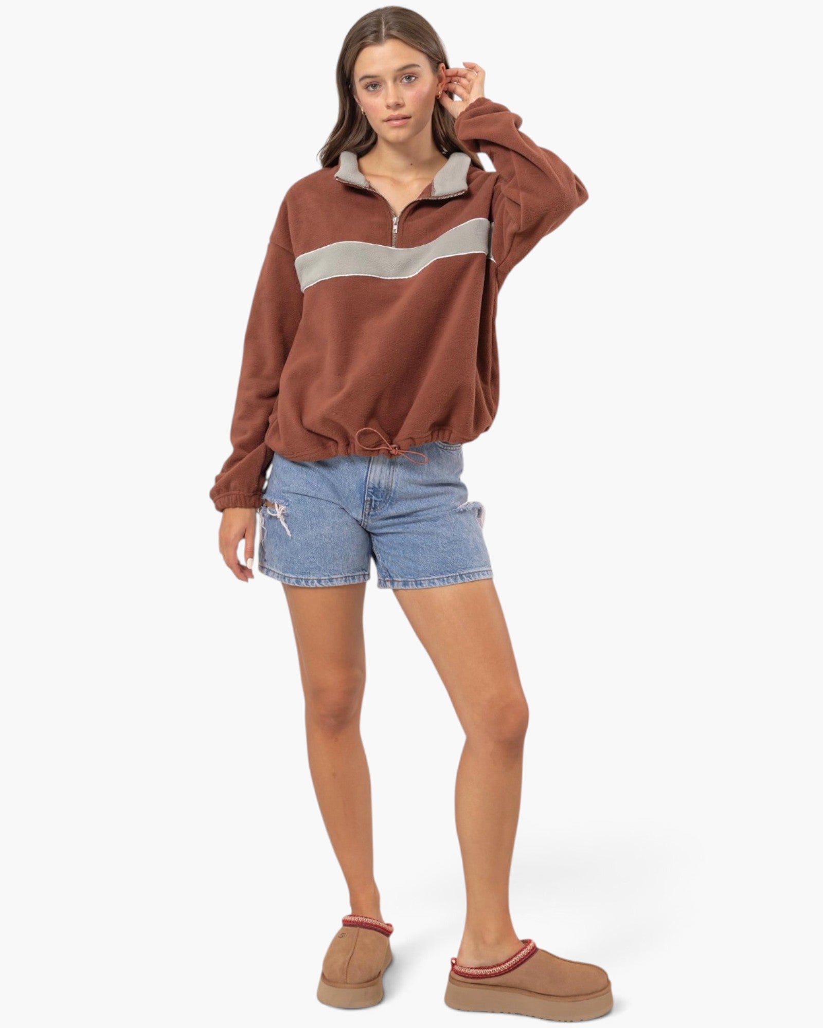 A woman modeling a brown half-zip sweatshirt with a light gray chest stripe, paired with denim shorts and tan slip-on shoes, against a plain white studio background.