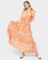 A woman modeling a flowing off-the-shoulder maxi dress with a vibrant orange, peach, and white bohemian floral pattern, paired with white slide sandals, set against a solid light gray background.