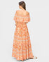A woman shown from the back wearing a long off-the-shoulder maxi dress with a colorful bohemian-style floral and geometric pattern in orange, peach, and cream, paired with white slide sandals, against a light gray studio background.