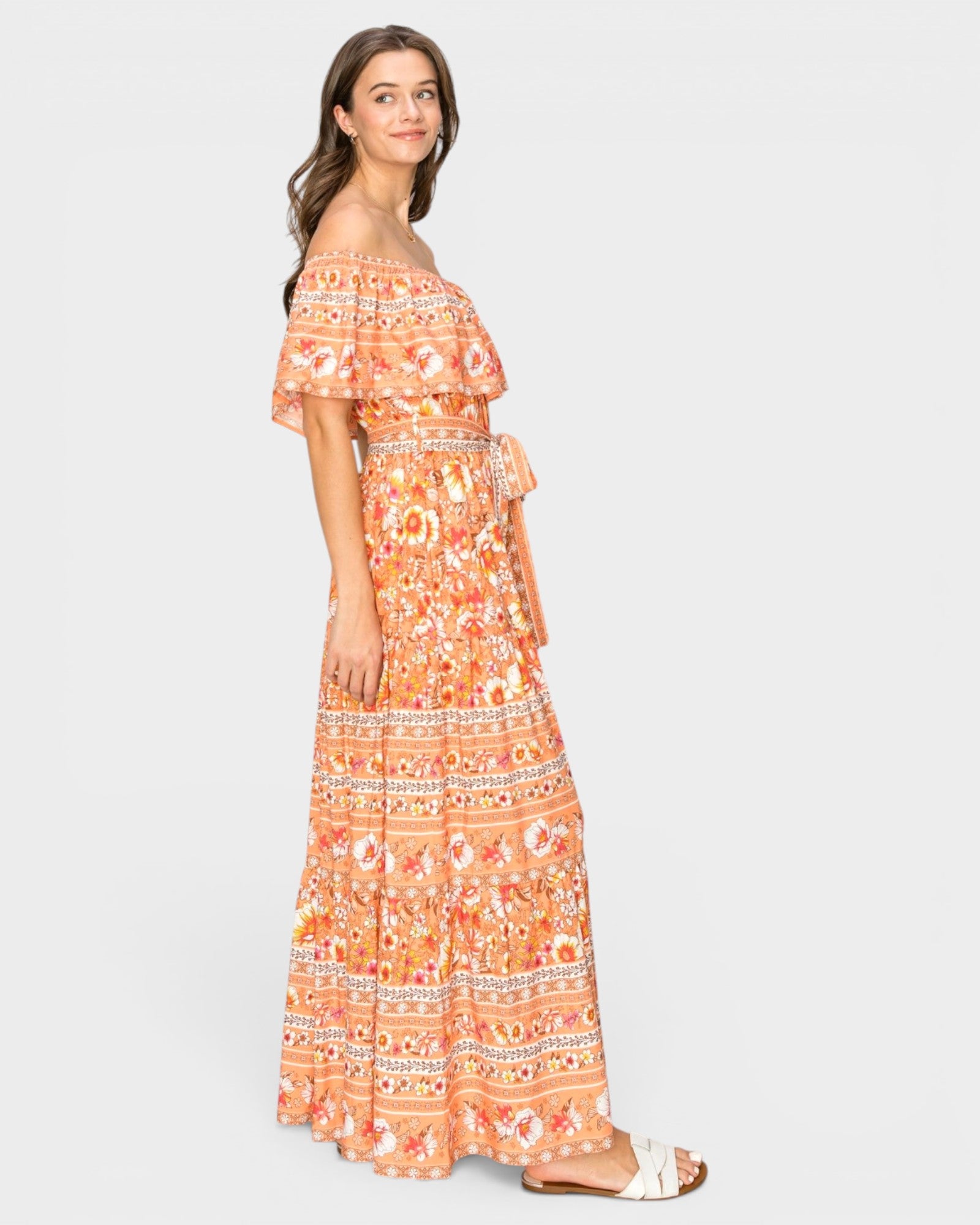 A woman modeling a long off-the-shoulder maxi dress with a vibrant orange, peach, and white bohemian-style floral pattern, shown against a solid light gray background.