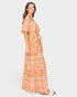 A woman modeling a long off-the-shoulder maxi dress with a vibrant orange, peach, and white bohemian-style floral pattern, shown against a solid light gray background.
