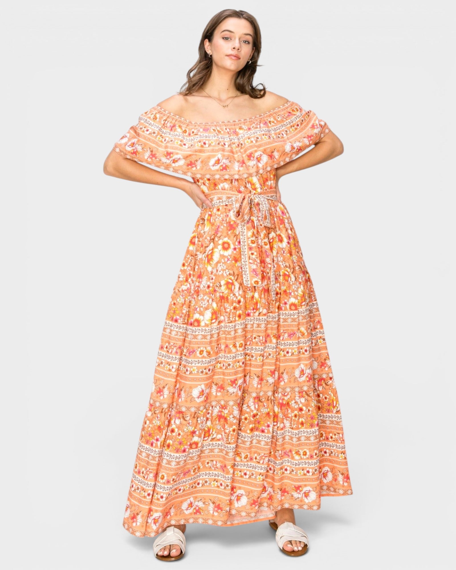 A woman modeling an off-the-shoulder maxi dress with a vibrant orange, peach, and white bohemian-style floral pattern, set against a solid light gray background.