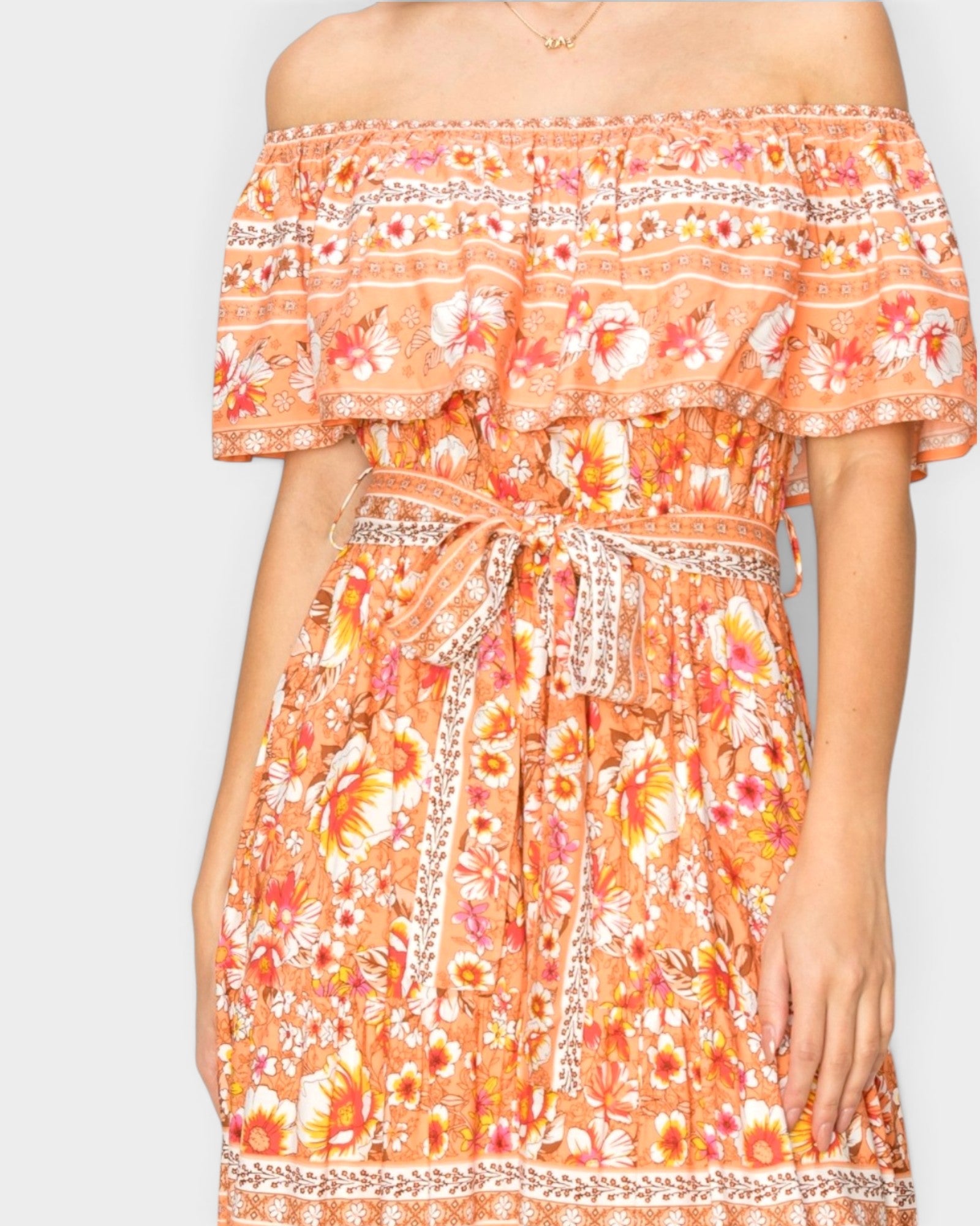 An off-the-shoulder orange floral dress with a ruffled neckline, a matching fabric tie belt, and intricate silver patterned trim, shown on a model against a light gray background.