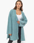A woman modeling a fuzzy, open-front cardigan in a dusty teal blue color, worn over a white tank top and black pants against a plain white background.