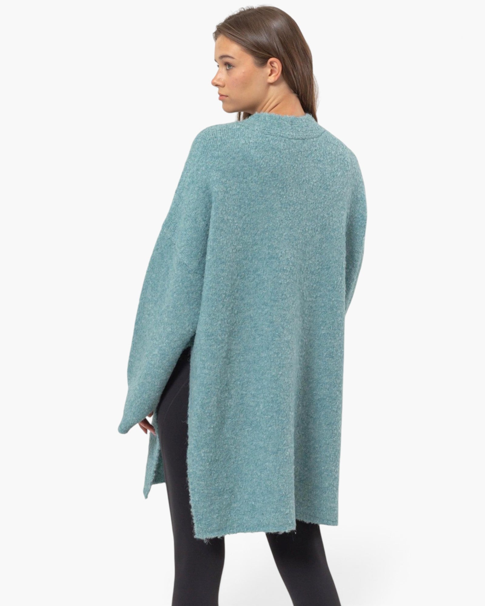 A woman wearing a fuzzy, textured knit poncho sweater in a dusty teal blue color, shown from a side-back angle against a plain white studio background.