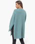 A woman wearing a fuzzy, textured knit poncho sweater in a dusty teal blue color, shown from a side-back angle against a plain white studio background.