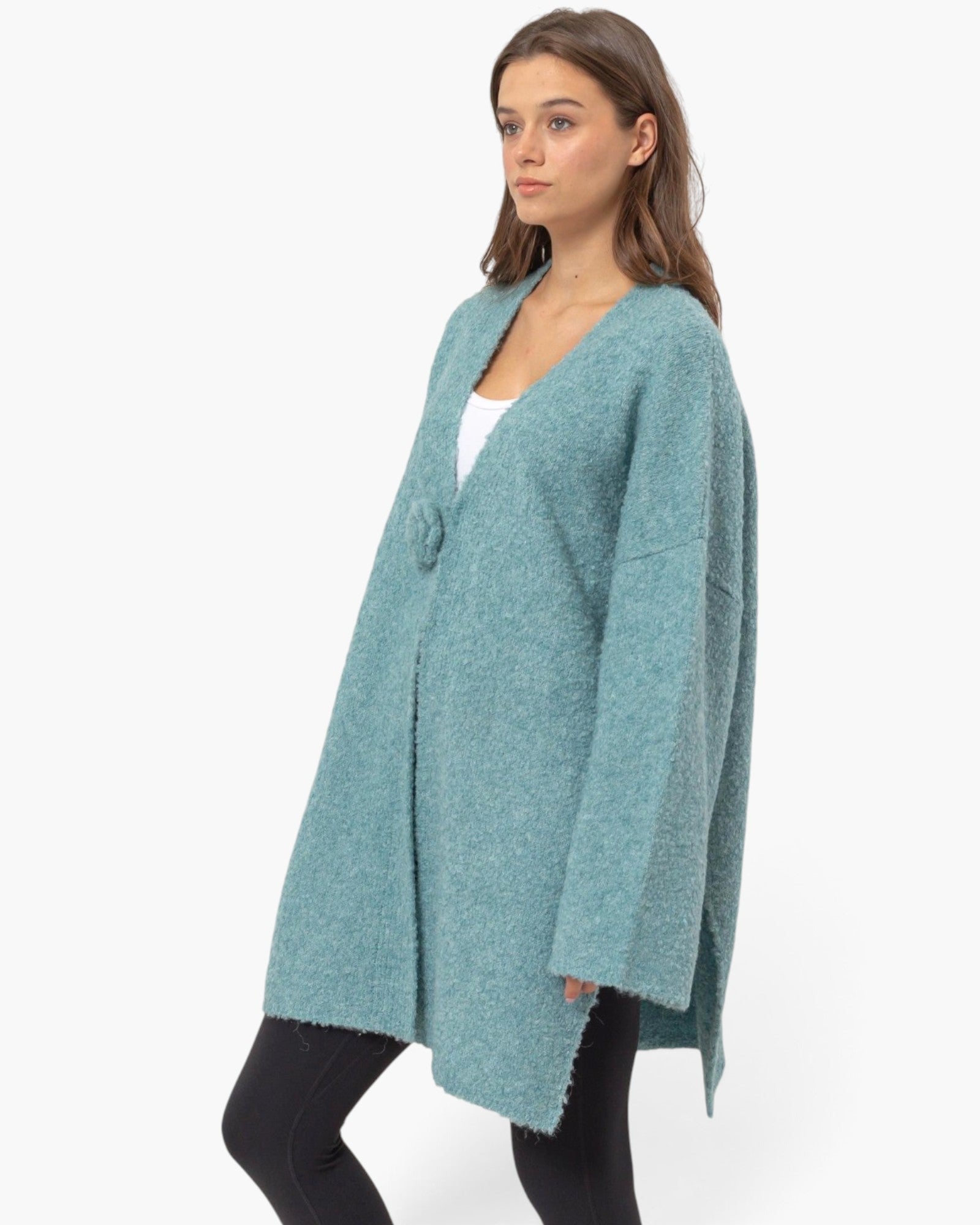 A woman modeling a long, open-front knit cardigan in a heathered teal blue color, styled over a white top and black leggings against a plain white studio background.