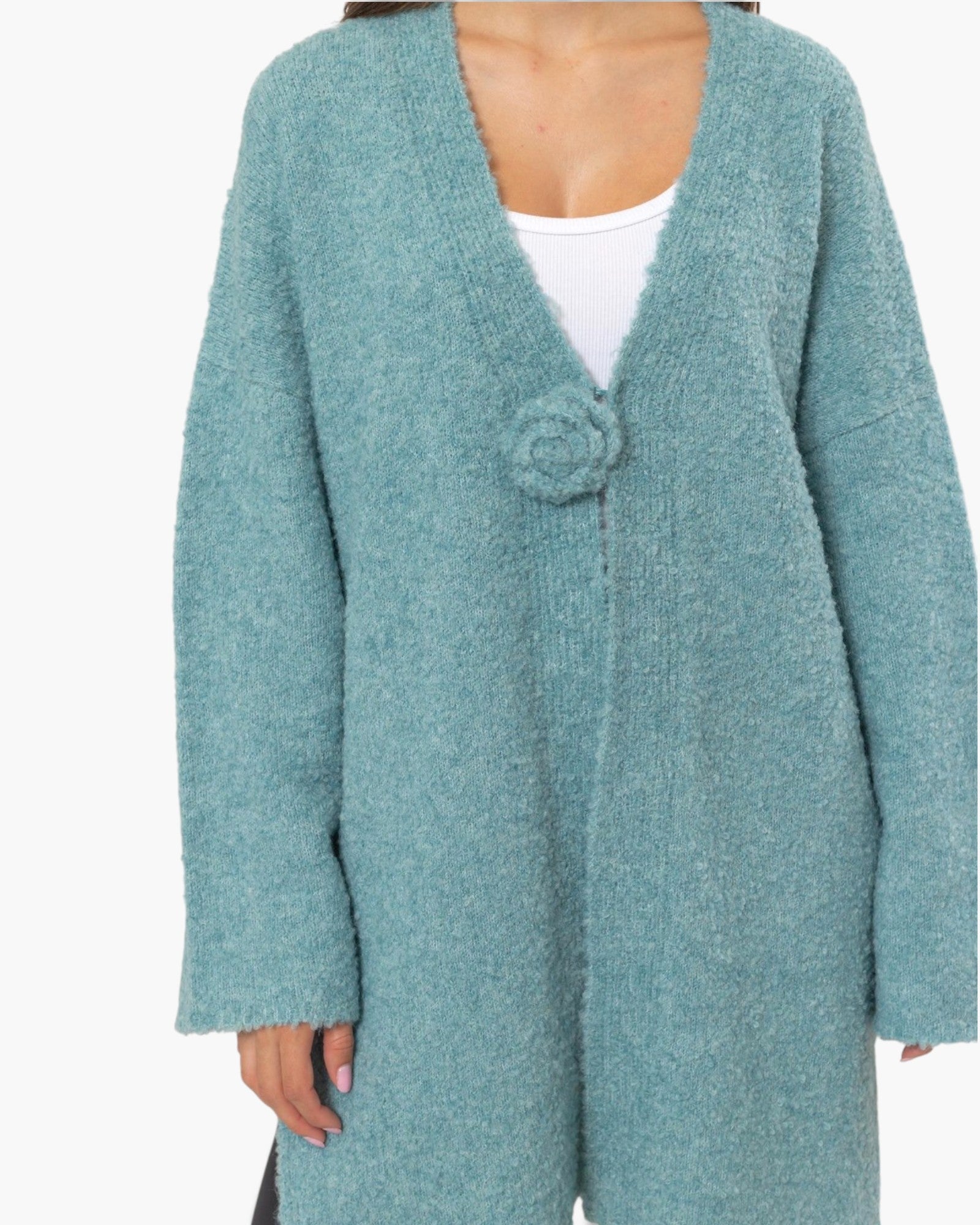 A person wearing a fuzzy, oversized knit cardigan in a dusty teal blue color. The cardigan features a large, decorative fabric flower embellishment on the chest, a deep V-neckline, and a white tank top underneath.