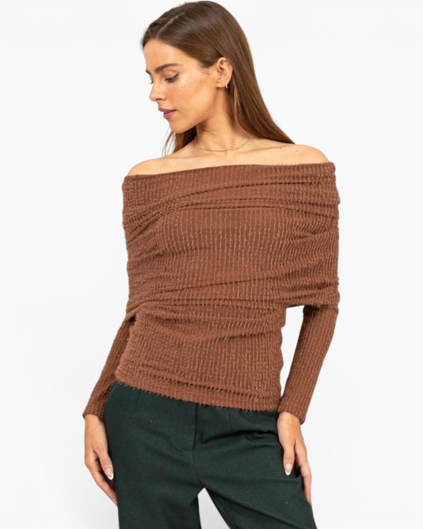 A woman wearing a brown off-the-shoulder ribbed knit sweater and dark green pants, photographed against a plain light gray studio background.