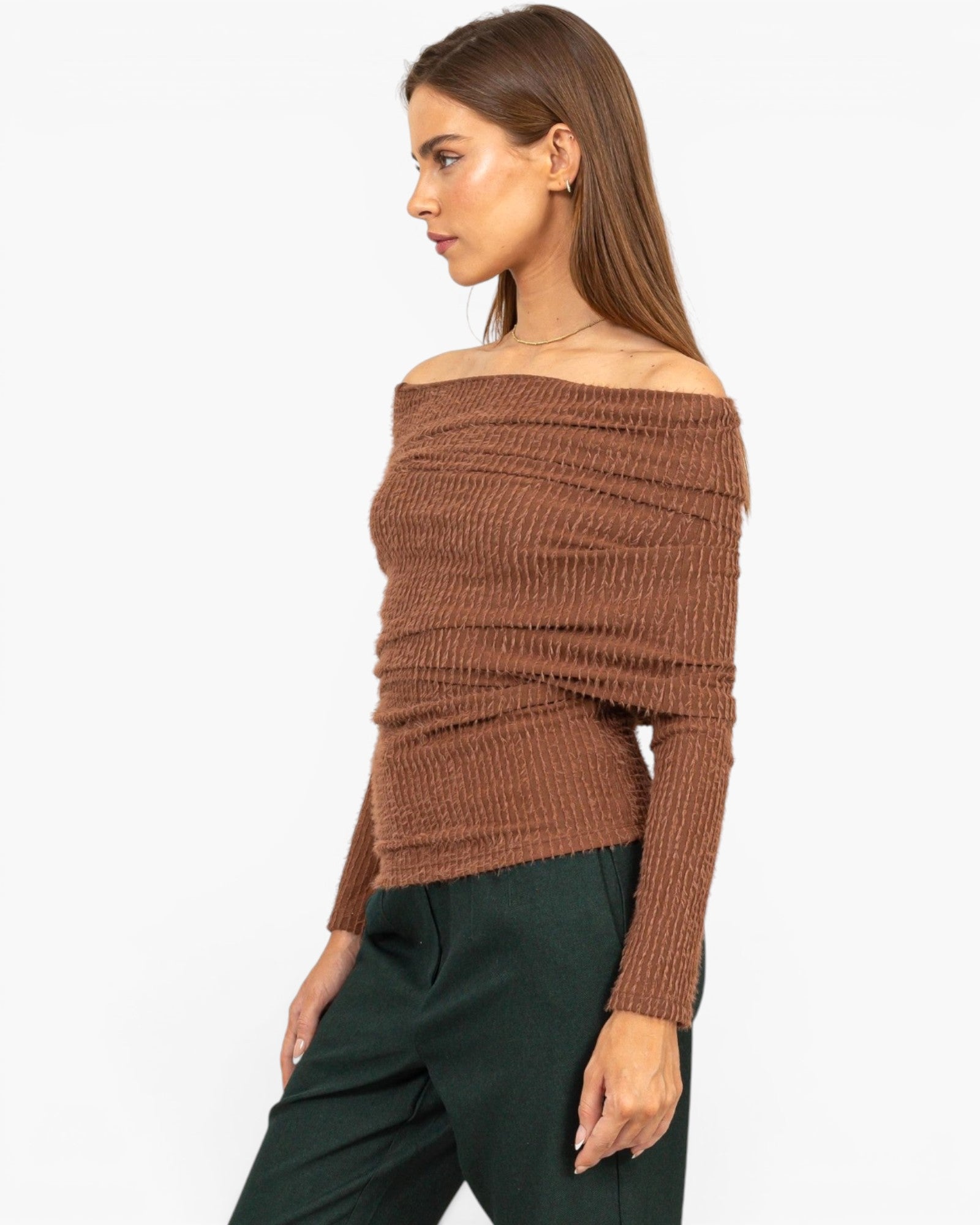 A woman wearing a brown off-the-shoulder ribbed knit sweater and dark green pants, shown in profile against a plain light gray studio background.
