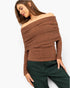 A woman wearing a brown off-the-shoulder ribbed knit sweater and dark green pants, photographed against a plain light background.