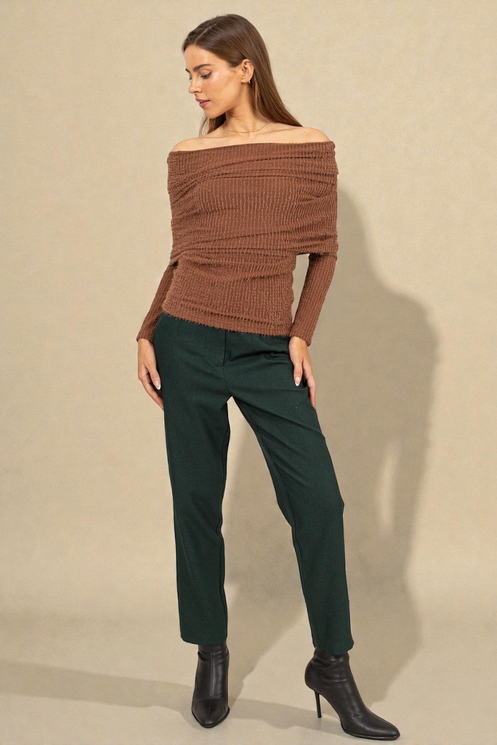 Woman wearing a brown off-shoulder sweater and green pants against a beige background
