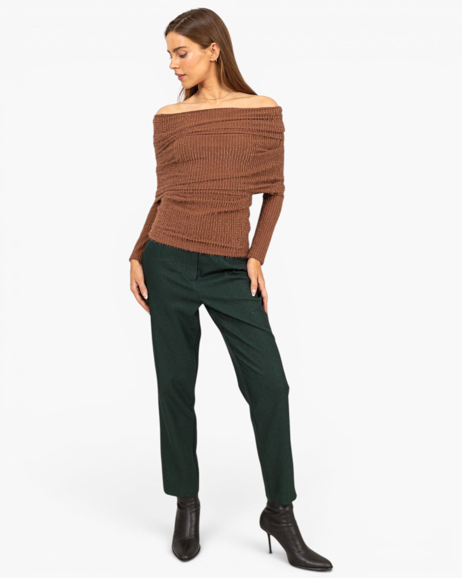 A woman modeling an off-the-shoulder brown ribbed knit sweater paired with dark green trousers and black ankle boots, photographed against a plain white studio background.