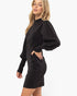 A woman modeling a black long-sleeve mini dress with a textured, sparkly finish and voluminous balloon sleeves, set against a plain white studio background.