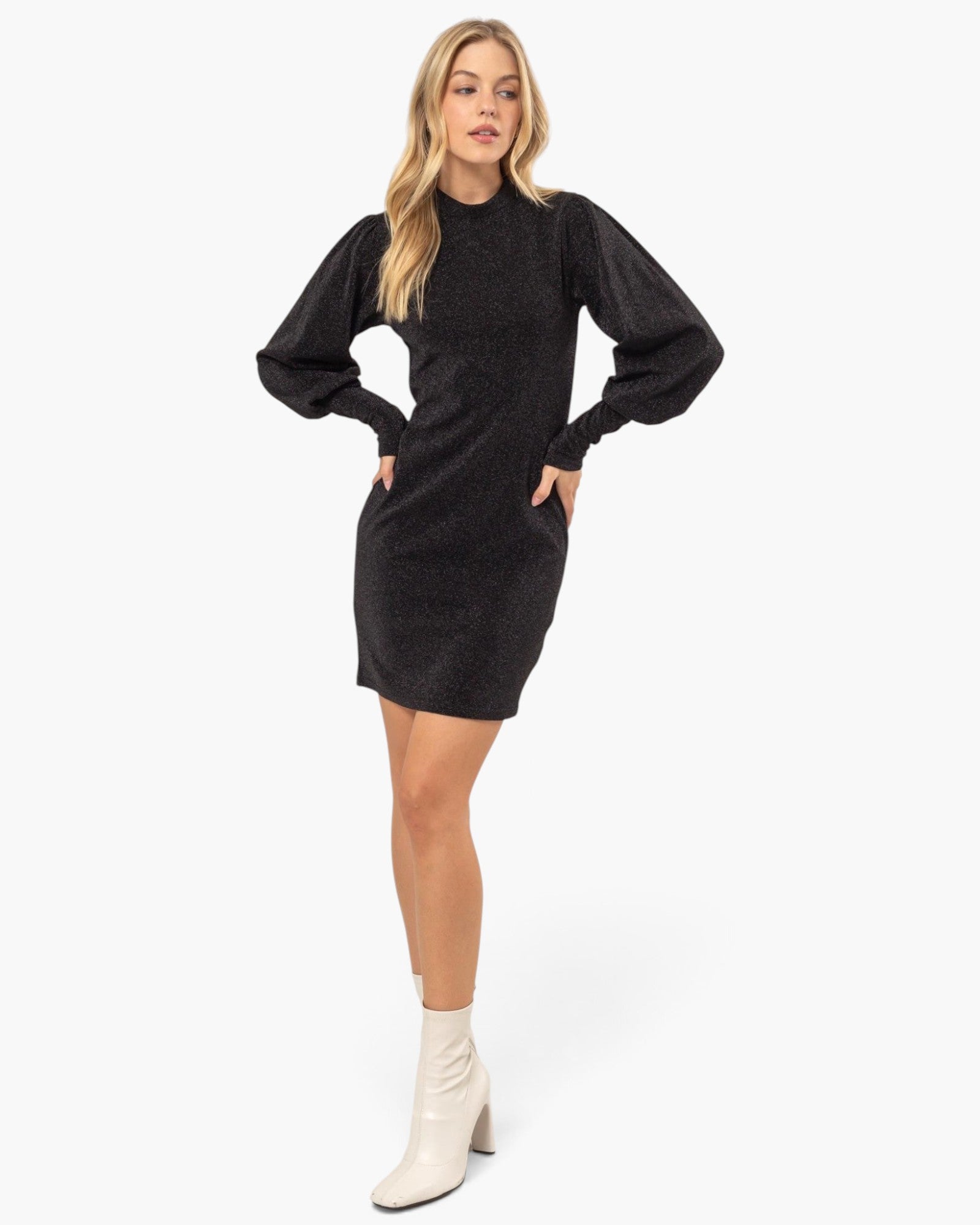 A woman modeling a black textured mini dress with voluminous balloon sleeves and white ankle boots, standing against a plain white studio background.