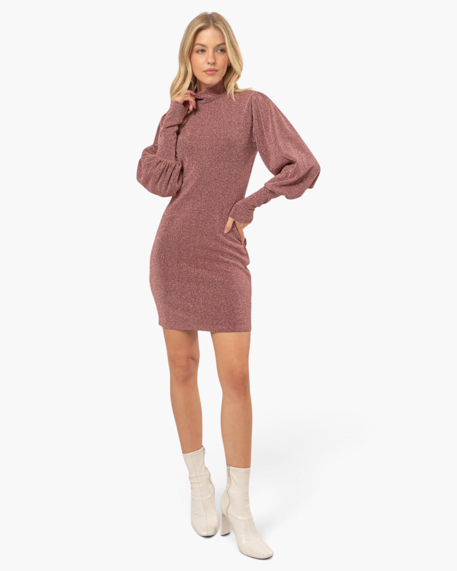 A woman modeling a dusty rose ribbed knit mini dress with balloon sleeves and a high mock neck, paired with off-white ankle boots, against a plain white studio background.