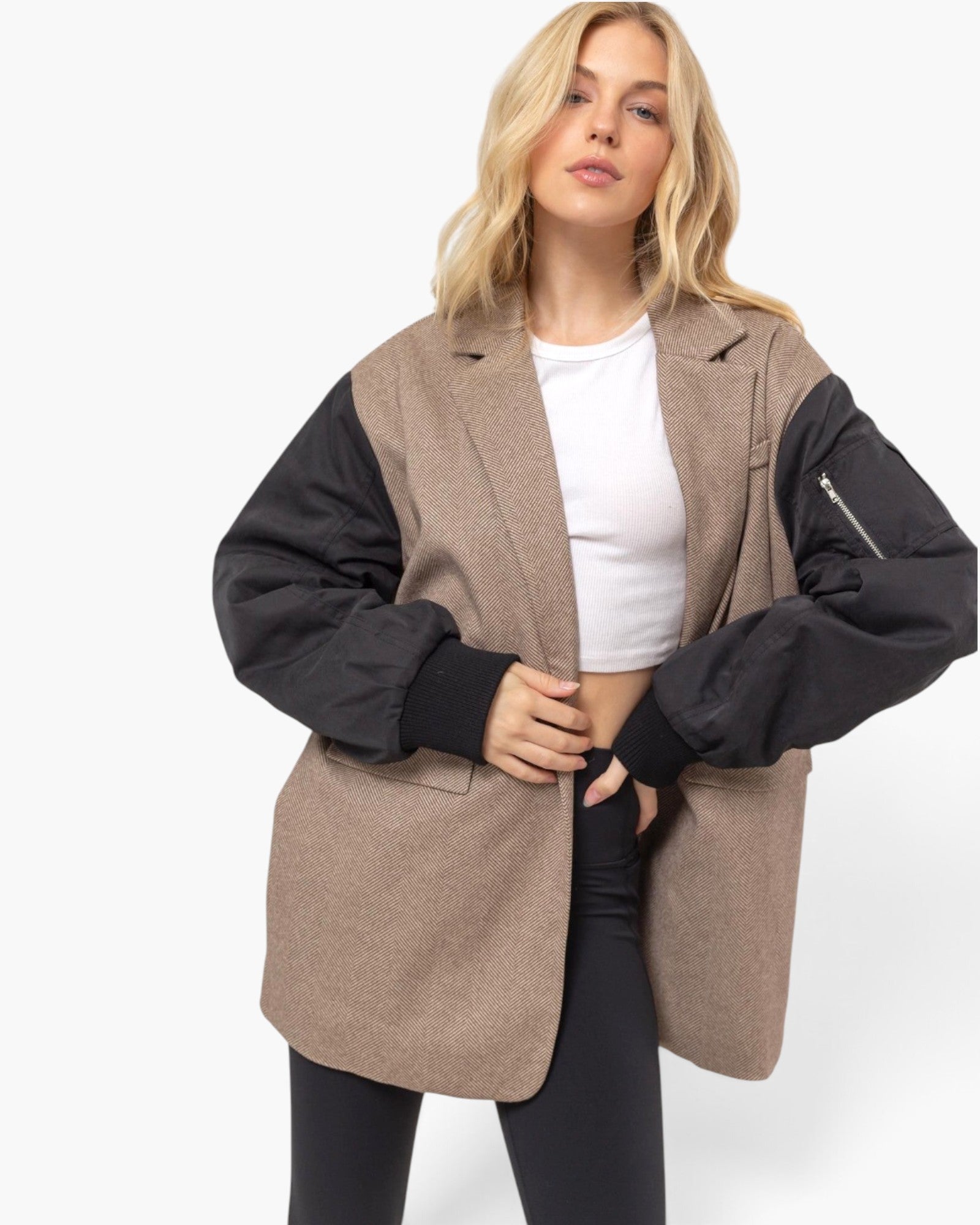 A woman modeling a color-blocked blazer vest with a taupe-brown body and black sleeves, worn over a white crop top and black leggings, against a plain white studio background.