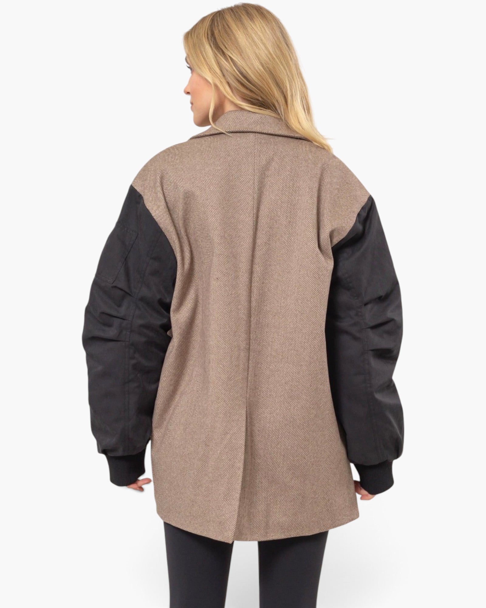 A woman shown from the back wearing a colorblock jacket with a taupe-brown body and black sleeves, paired with black leggings, against a plain white background.