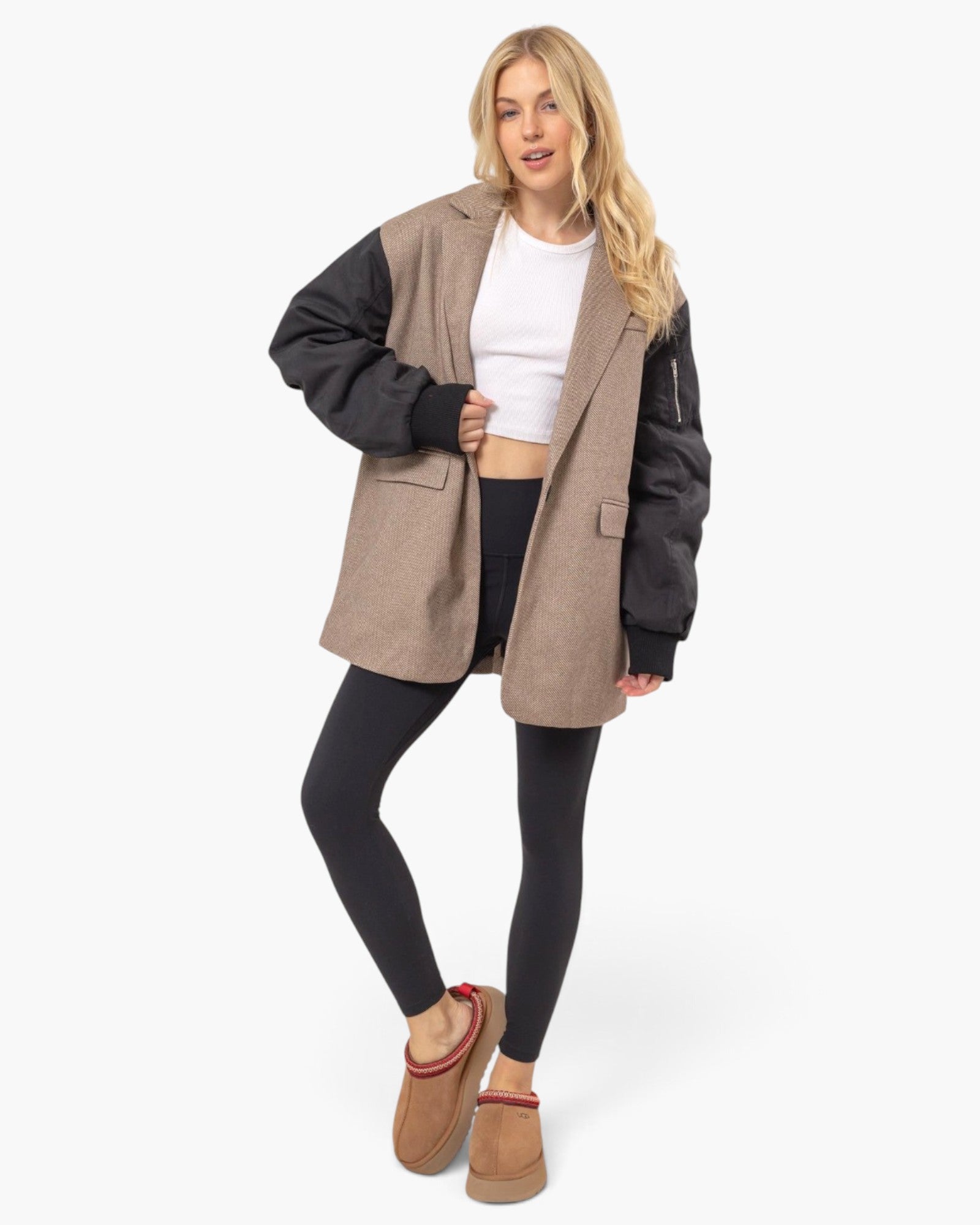 A woman modeling a colorblock blazer with taupe wool body and black sleeves, worn open over a white crop top and black leggings, paired with tan suede slippers against a light gray studio background.