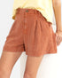 A close-up of a model wearing high-waisted, pleated shorts in a rust-orange color, paired with a light yellow top against a plain white background.