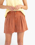 A model wearing high-waisted, pleated terracotta shorts with a yellow top, shown from the chest down against a plain white background.
