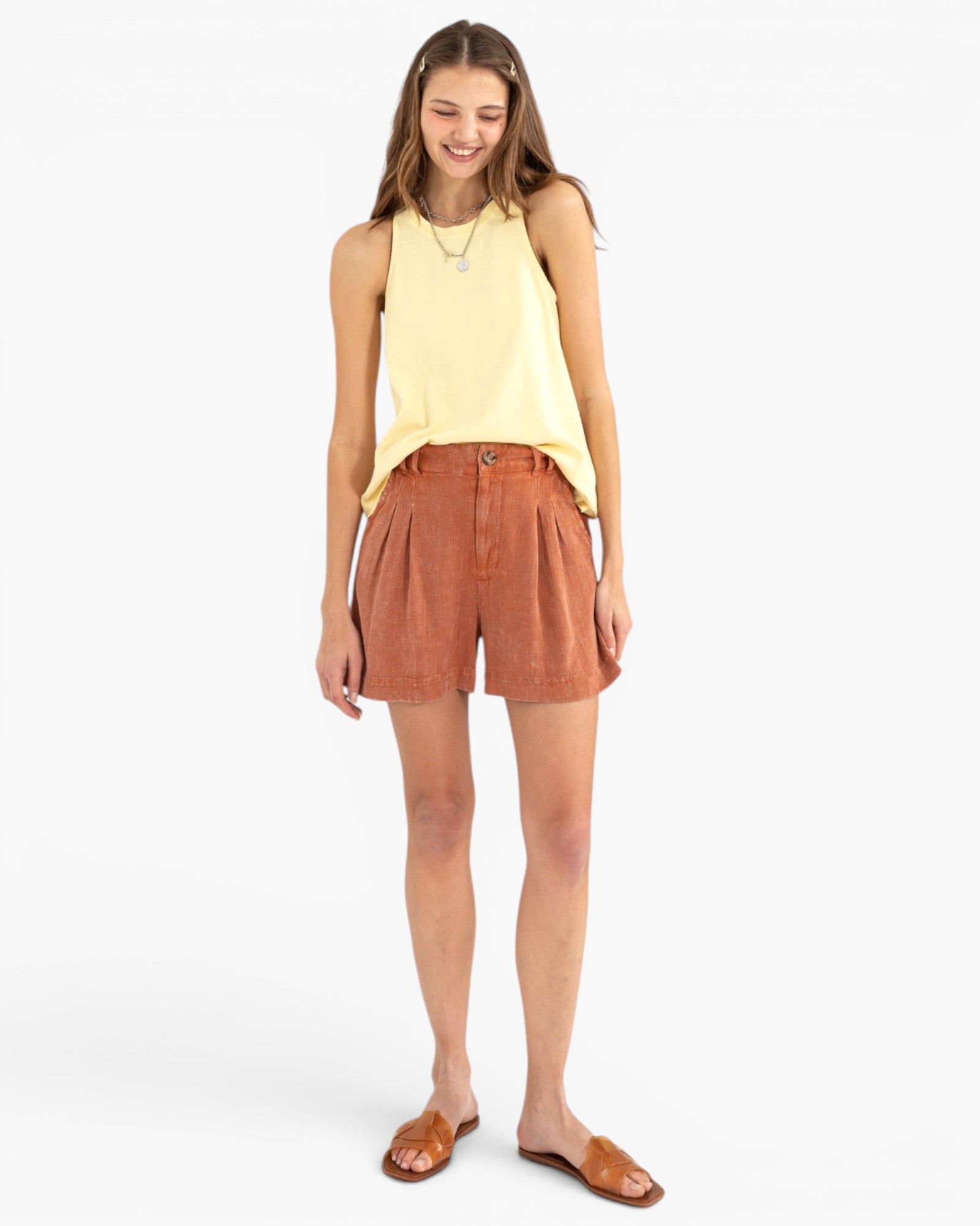 A woman modeling a light yellow sleeveless top and rust-orange pleated shorts against a plain light gray studio background.
