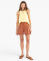 A woman modeling a light yellow sleeveless top and rust-orange pleated shorts against a plain light gray studio background.