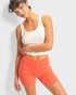 Woman wearing a white tank top with small patterns and bright orange shorts on a light gray background