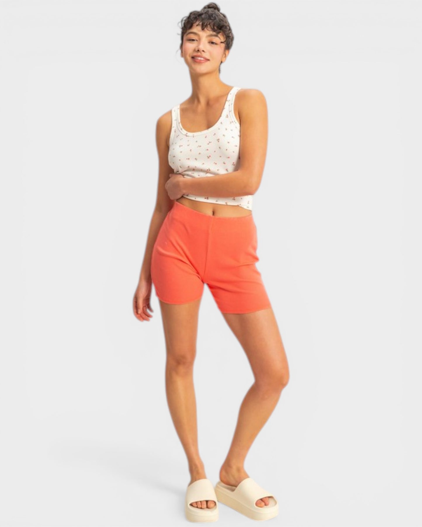Woman wearing a white sleeveless top and orange shorts on a light gray background