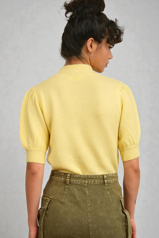 Person wearing a yellow cardigan and green pants against a gray background