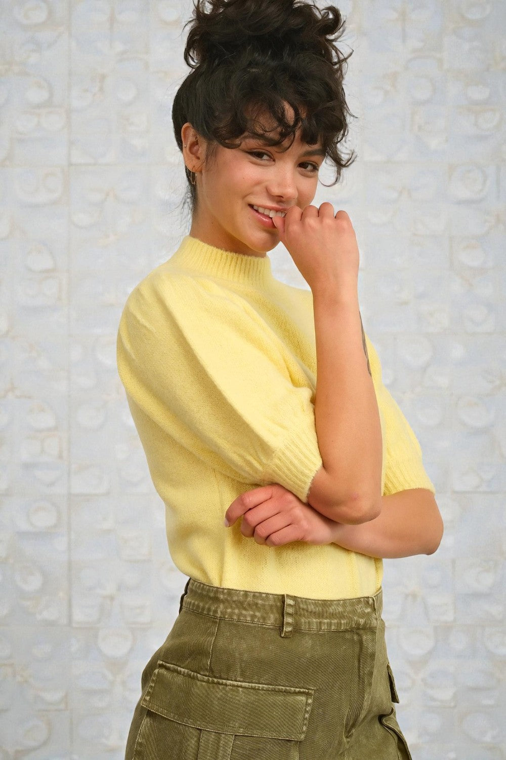 Woman wearing a yellow sweater and green pants against a light textured wall.