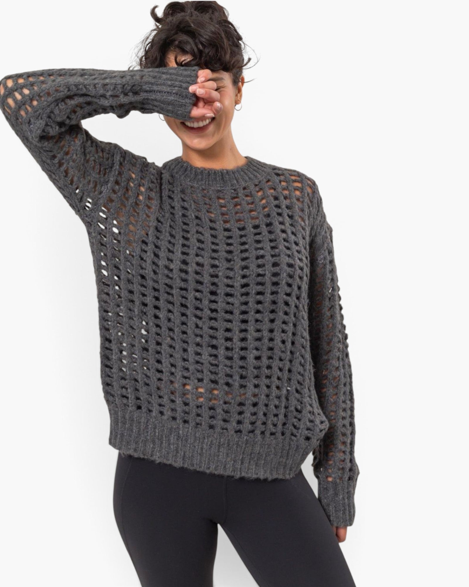 A woman wearing a dark gray open-knit crewneck sweater and black leggings, posing against a plain white background.