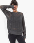 A woman wearing a dark gray open-knit crewneck sweater and black leggings, posing against a plain white background.