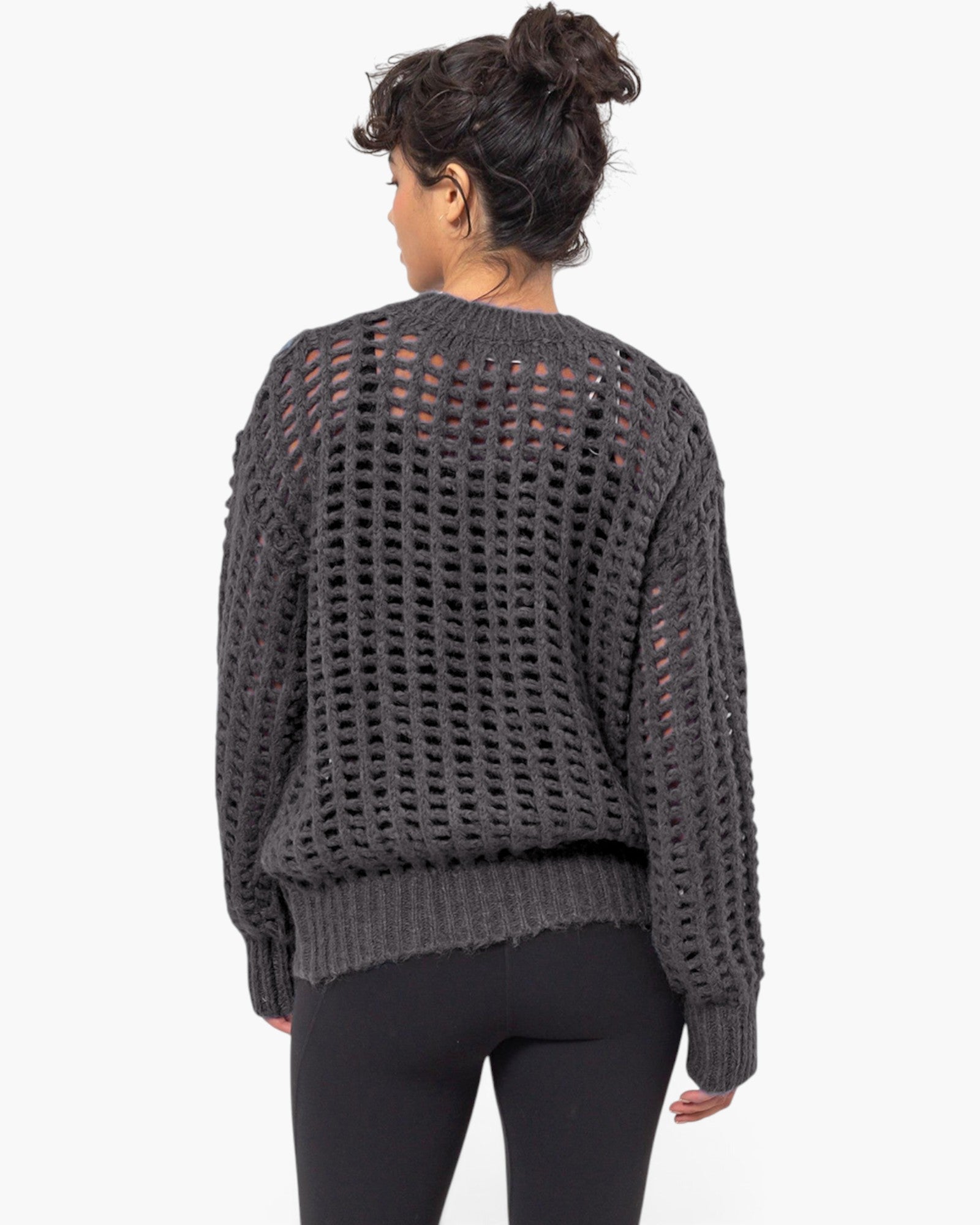 A woman viewed from the back wearing a dark charcoal gray open-knit sweater and black leggings against a plain white studio background.