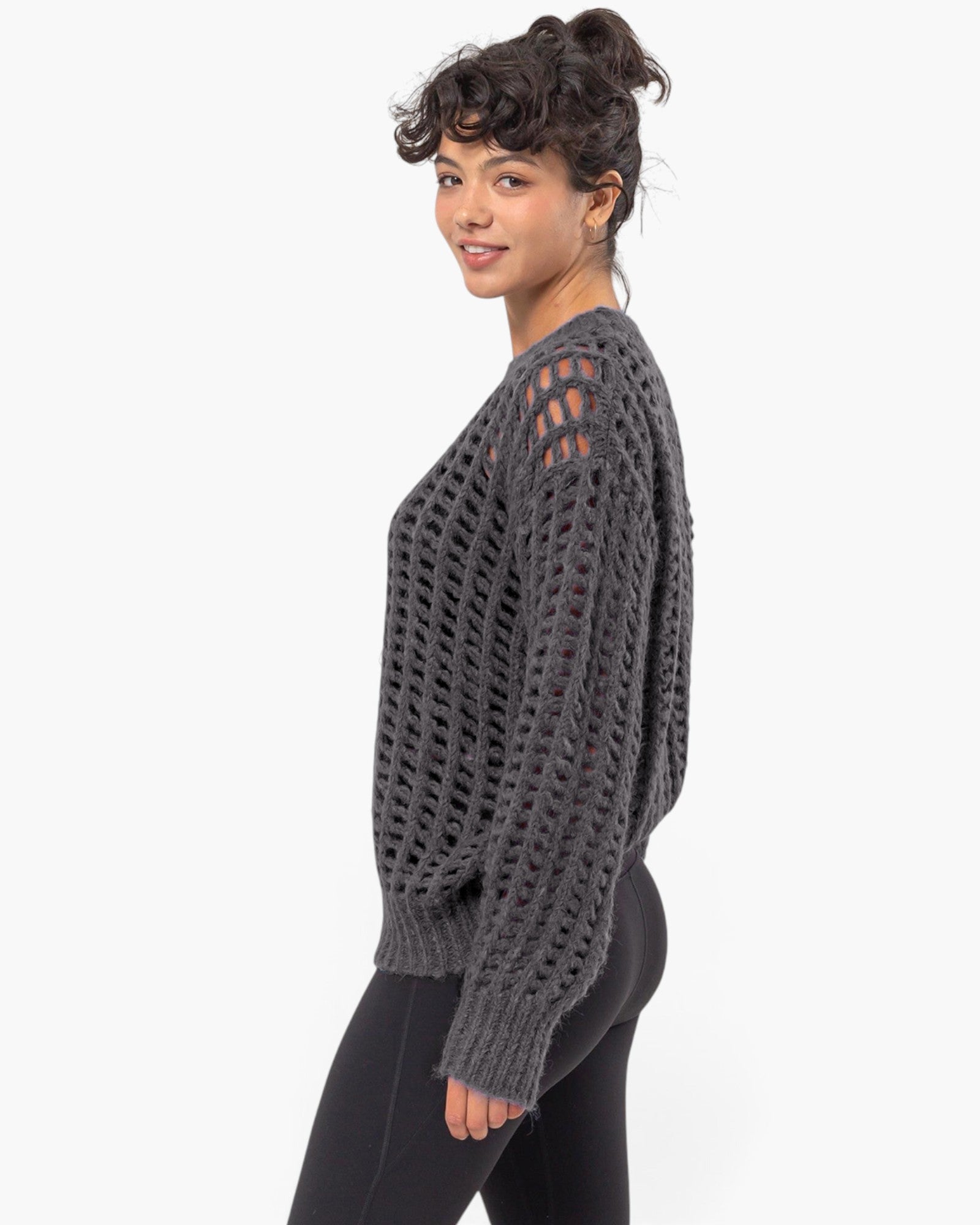 A woman modeling a dark gray open-knit sweater with side cutouts, paired with black leggings, against a plain white studio background.
