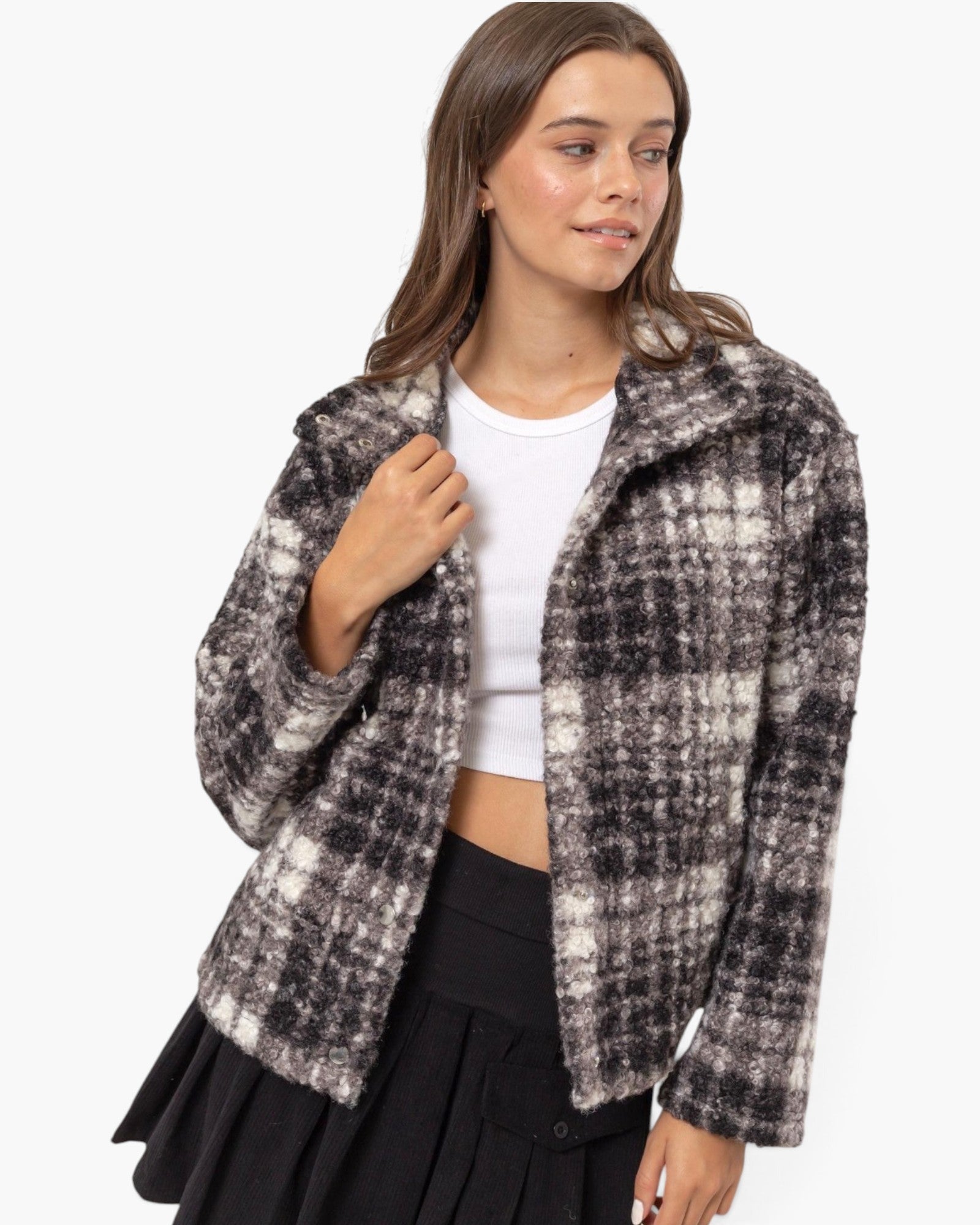 A woman modeling a fuzzy, cropped plaid jacket in black and off-white, worn over a white crop top and a black pleated skirt against a plain white background.