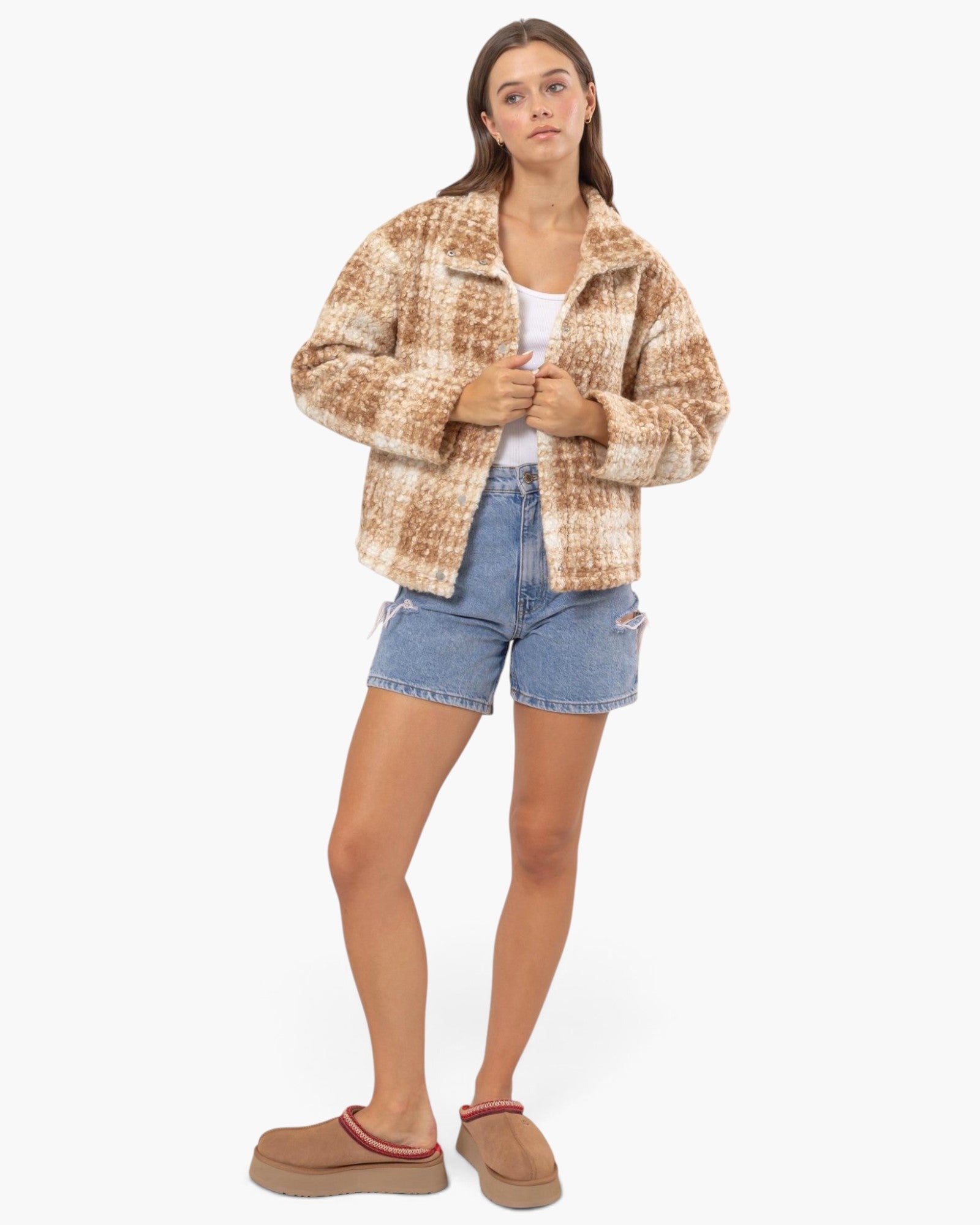 A woman modeling a cropped, fuzzy sherpa-style jacket in a tan and brown marled pattern, paired with a white tank top, distressed denim shorts, and brown suede slip-on mules, against a plain white studio background.
