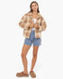 A woman modeling a cropped, fuzzy sherpa-style jacket in a tan and brown marled pattern, paired with a white tank top, distressed denim shorts, and brown suede slip-on mules, against a plain white studio background.