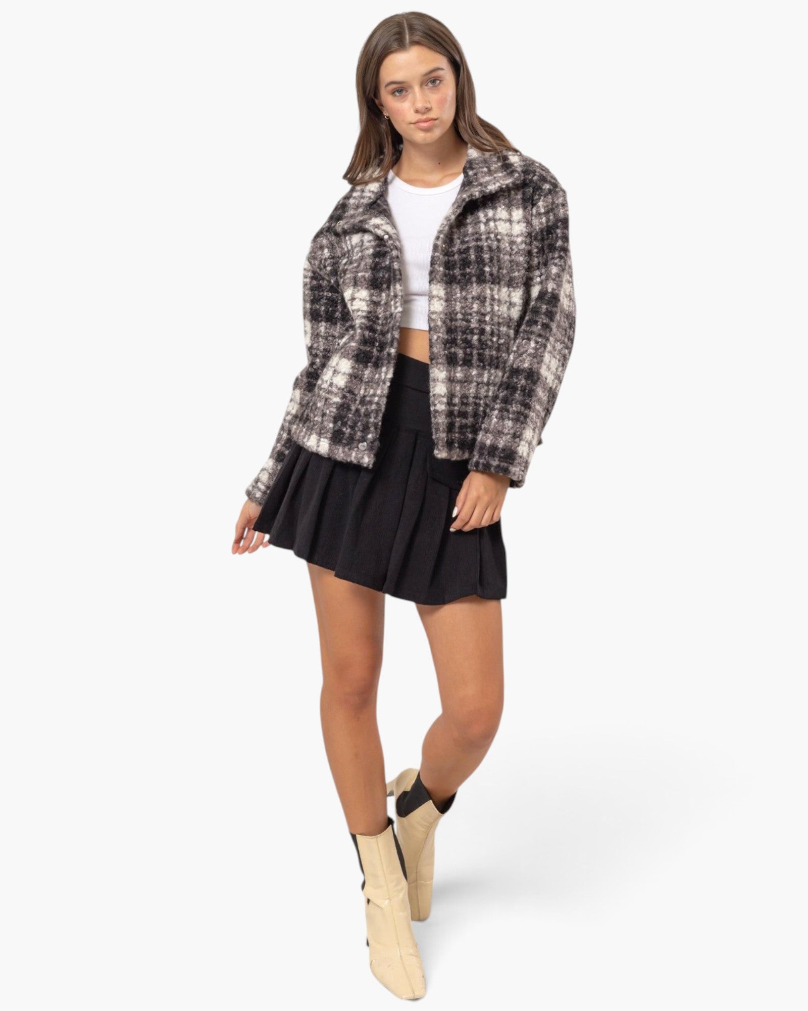A model wearing a black and white plaid cropped jacket, a black pleated mini skirt, a white crop top, and beige ankle boots, standing against a light gray studio background.
