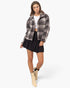A model wearing a black and white plaid cropped jacket, a black pleated mini skirt, a white crop top, and beige ankle boots, standing against a light gray studio background.