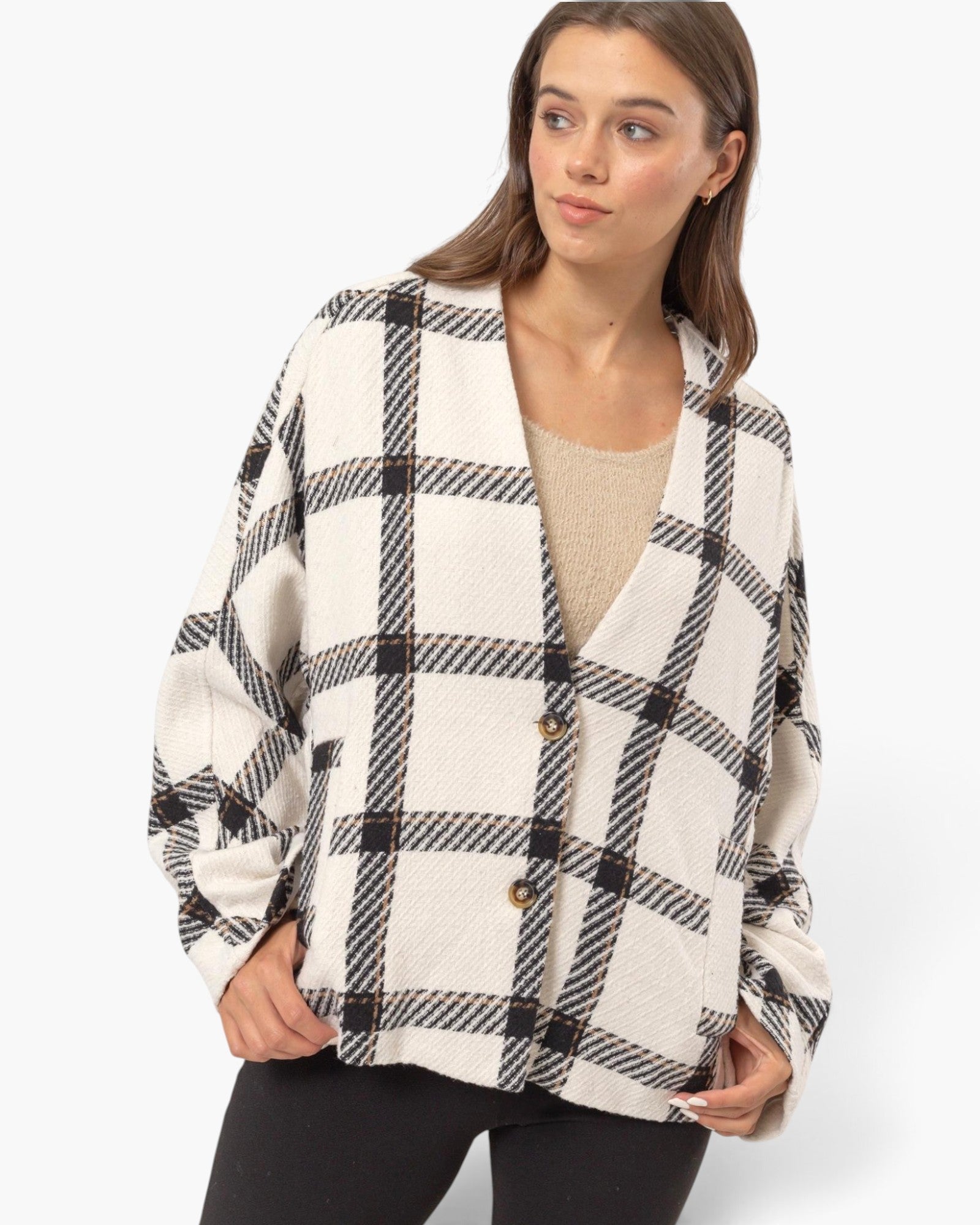 A woman wearing a cream-colored knit cardigan with a black and gray plaid pattern, featuring three brown buttons, over a beige top and black pants, set against a plain light gray background.