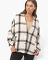 A woman wearing a cream-colored knit cardigan with a black and gray plaid pattern, featuring three brown buttons, over a beige top and black pants, set against a plain light gray background.