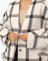 A close-up of a person wearing an oversized white plaid coat with black and gray diagonal stripes, layered over a beige knit top. The coat features a single visible brown button and a textured fabric.