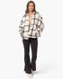 A woman modeling a black and off-white plaid button-up jacket with oversized sleeves, paired with black flared pants and off-white boots, against a plain white studio background.