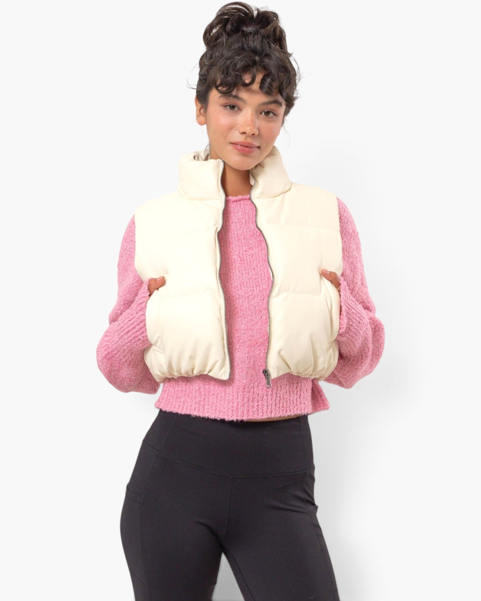A woman modeling a cream puffer vest over a pink knit sweater and black leggings, photographed against a clean white studio background.